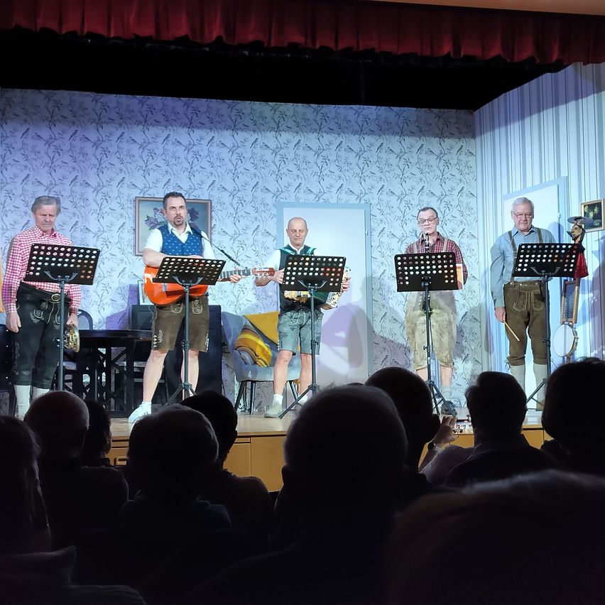 Five men on stage play instruments in front of an audience. The central figure plays a guitar. The stage is set with musical stands and a backdrop of wallpaper.