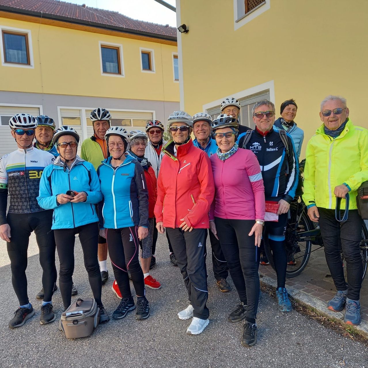 Eine Gruppe von Radfahrern in Fahrradausrüstung, einschließlich Helmen, Jacken und Brillen, steht vor einem gelben Gebäude mit mehreren Fenstern. Einige halten Fahrräder, andere posieren für ein Foto.