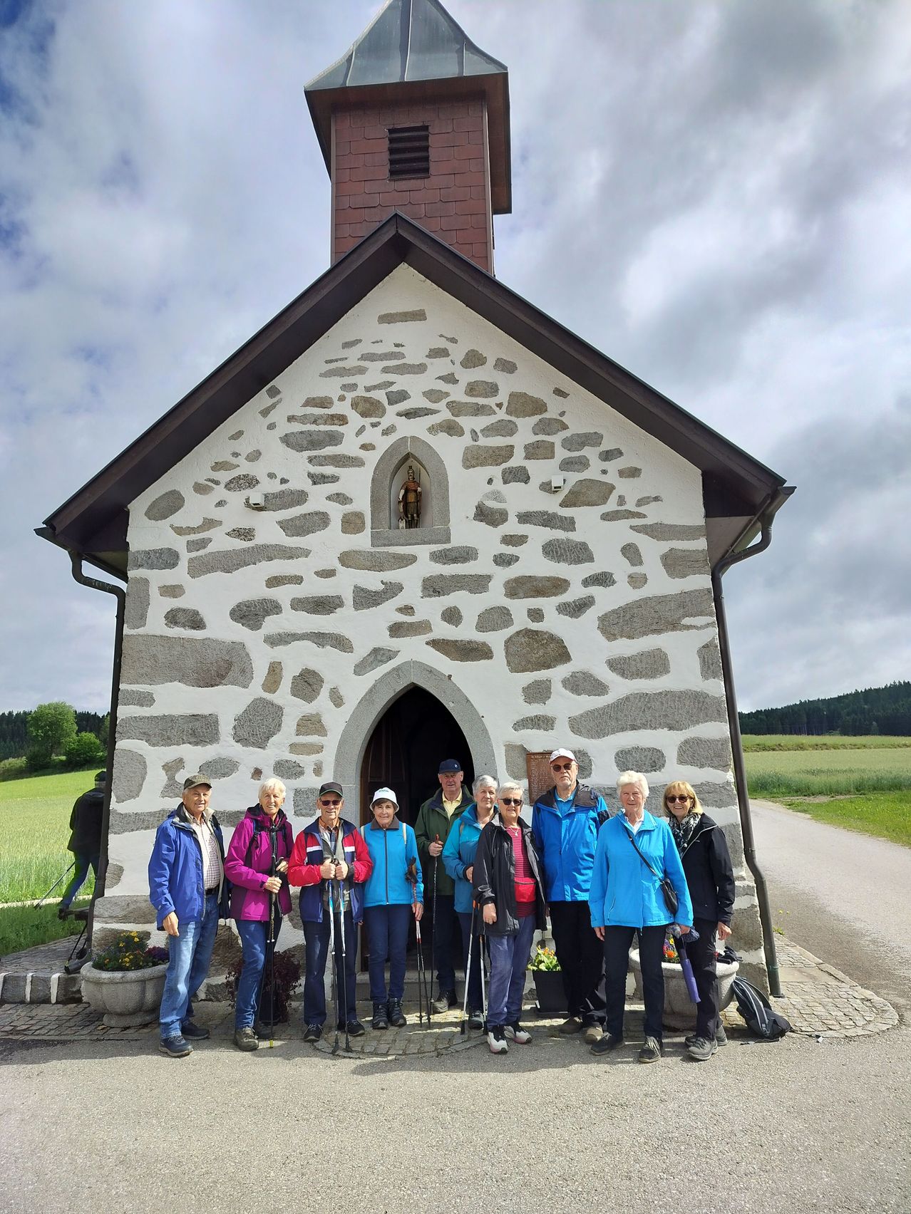 Eine Gruppe älterer Wanderer, einige mit Brille, steht vor einer Steinkapelle und hält Wanderstöcke in der Hand. Die Kapelle ist von einem grünen Feld umgeben, mit einer Person, die im Hintergrund geht.