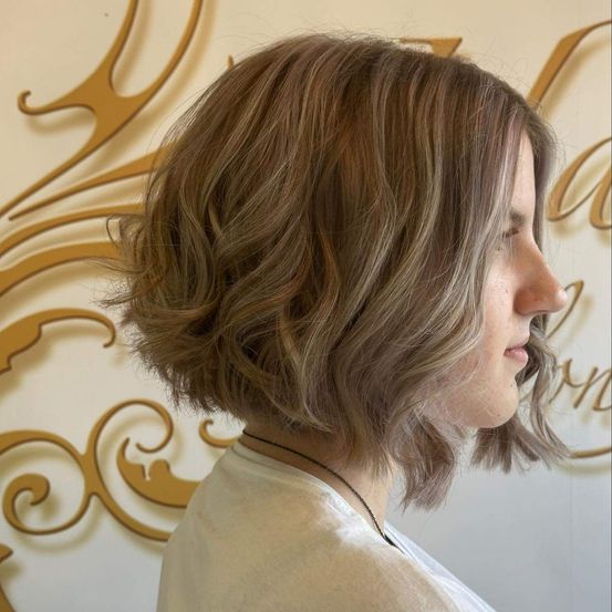 Eine Frau mit kurzen, welligen blonden Haaren und einem weißen Shirt schaut zur Seite. Sie steht vor einer Wand mit goldenen Mustern.
