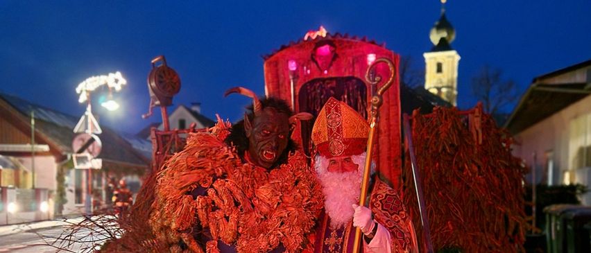 Zwei Personen in Kostümen stehen nachts auf einer Kopfsteinpflasterstraße. Einer ist ein Teufel mit rotem Outfit und Hörnern, der andere ein Bischof mit rotem Gewand und Hut. Sie stehen in der Nähe einer dekorativen Kutsche mit rotem Stoff.