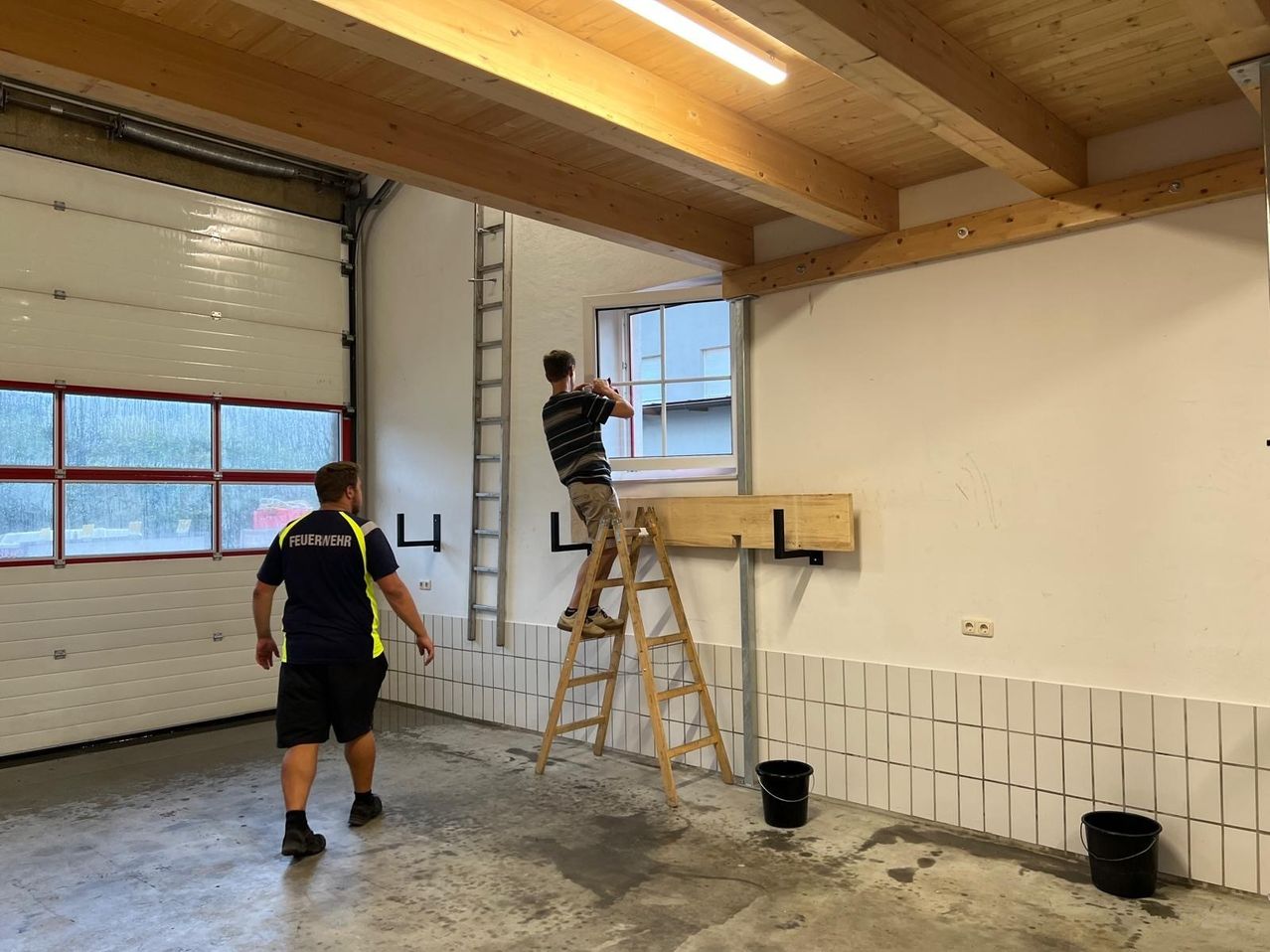Zwei Männer arbeiten in einer Garage. Einer ist auf einer Leiter und reinigt ein Fenster, während der andere zusieht. An der Wand stehen Eimer und eine Leiter.