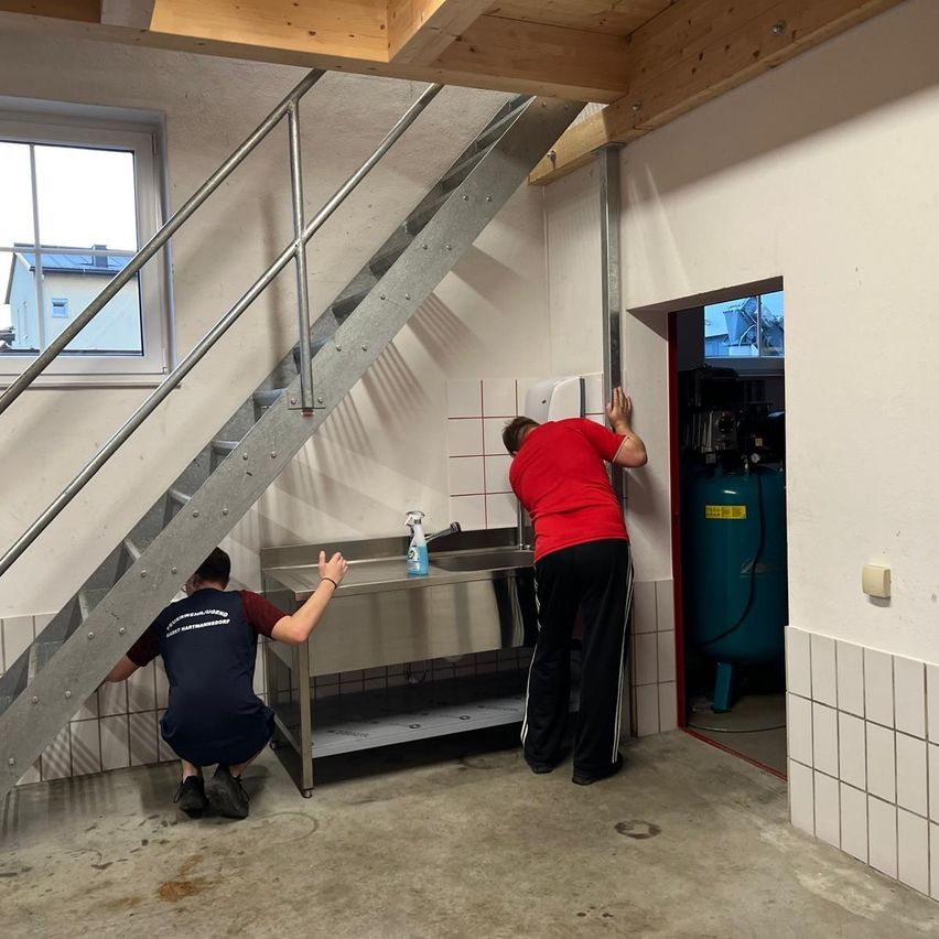 Zwei Personen befinden sich in einem Keller, eine kniet und die andere steht. Ein Waschbecken und eine Treppe sind in der Nähe.