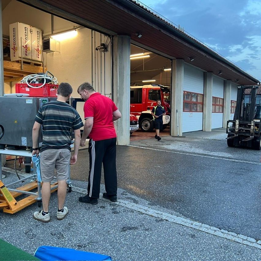 Zwei Männer stehen vor einer Feuerwache, ein Gabelstapler und ein Feuerwehrwagen sind im Inneren zu sehen.