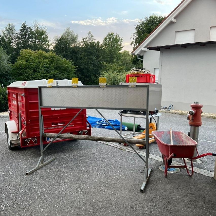 Ein Metallständer steht vor einem roten Anhänger mit weißem Dach. Ein Schubkarren befindet sich neben einem Feuerhydranten auf der Straße. Im Hintergrund ist ein Haus mit Schornstein zu sehen.