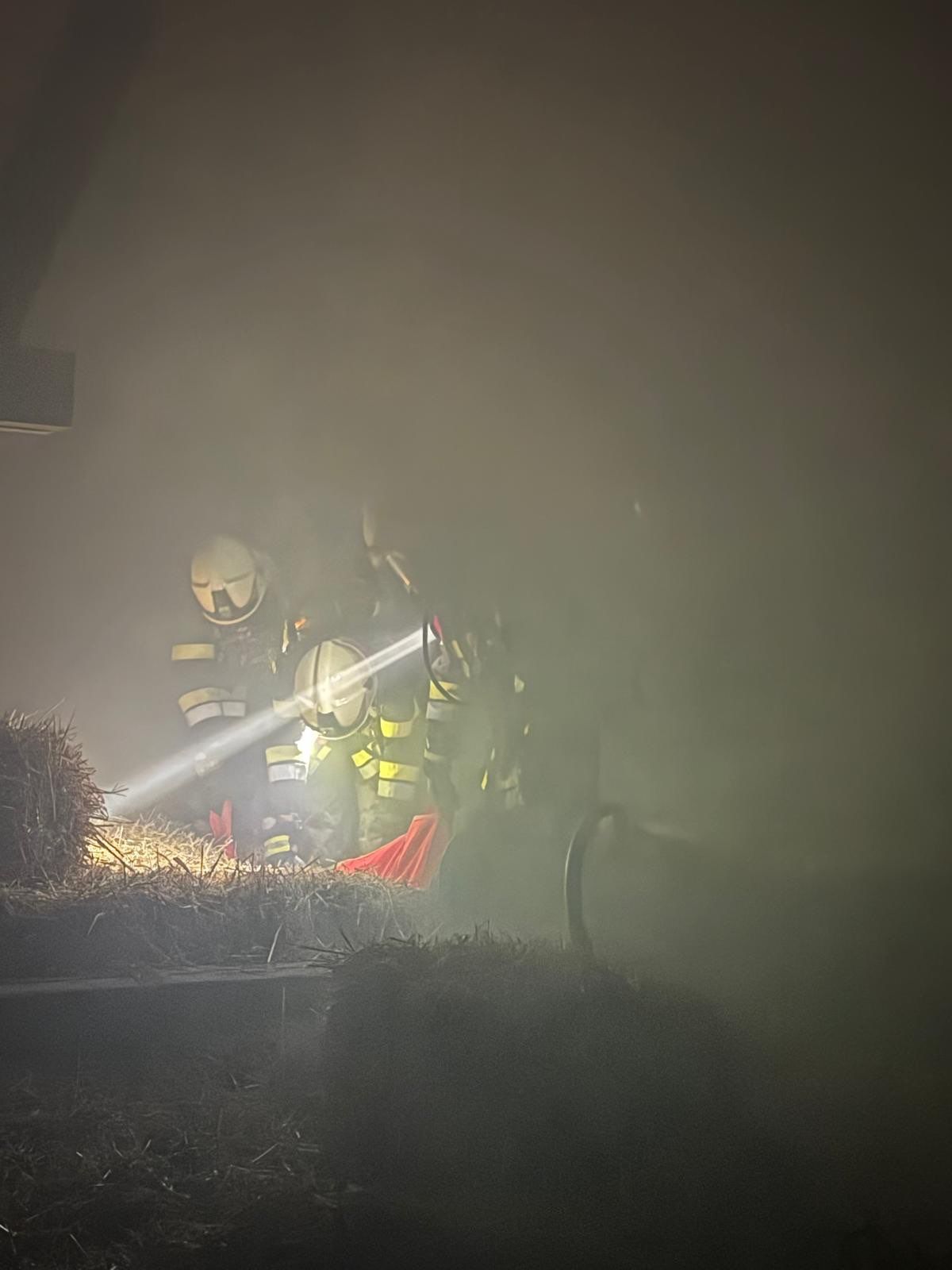 Feuerwehrleute in Helmen durchsuchen bei Nacht einen Heuballen mit einer Taschenlampe. Die Szene ist rauchig.