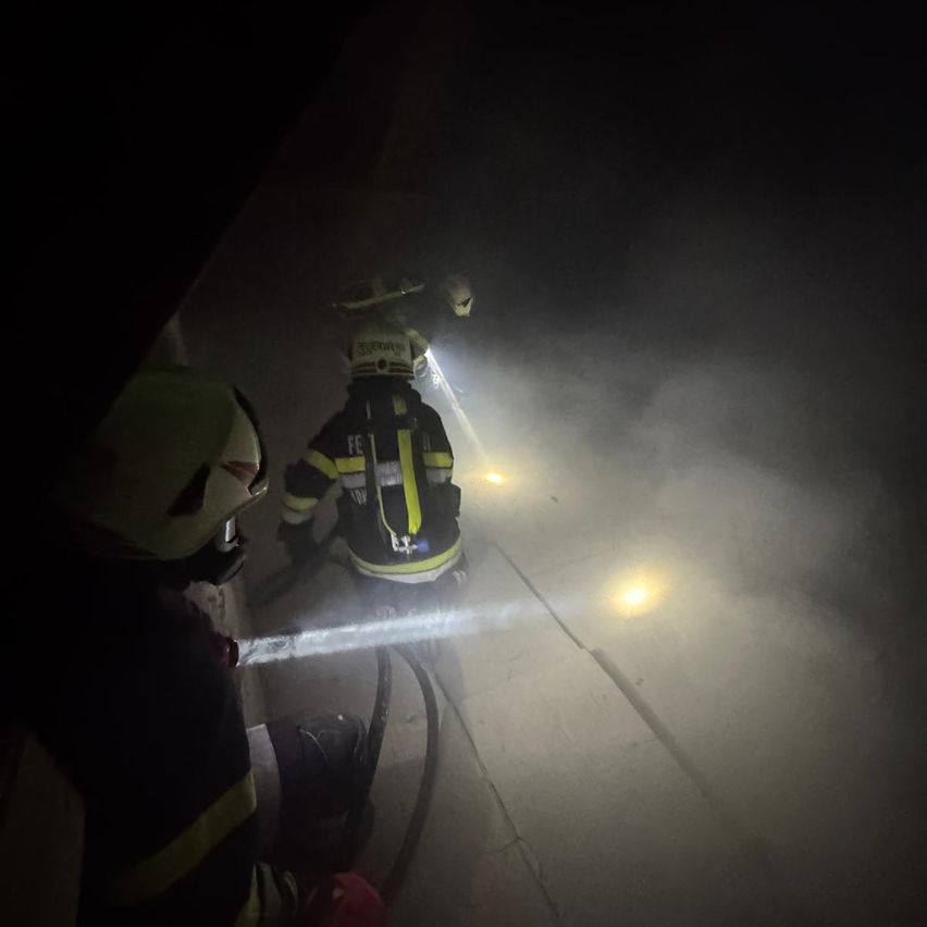 Zwei Feuerwehrleute in Schutzausrüstung, ausgestattet mit Helmen und Taschenlampen, navigieren durch einen rauchigen, schwach beleuchteten Tunnel. Einer hält einen Schlauch, während der andere ein Licht leuchtet.