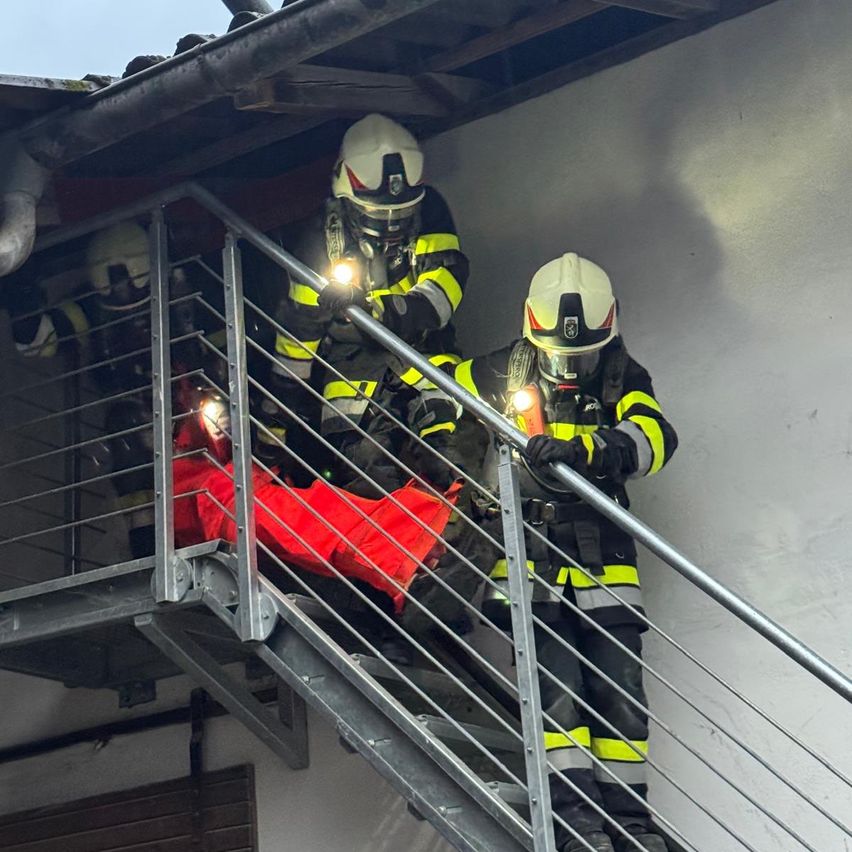Drei Feuerwehrleute klettern eine Metalltreppe hinunter und tragen Helme und Schutzanzüge. Sie halten ein rotes Objekt, möglicherweise eine Feuerwehraxt, und benutzen Taschenlampen. Einer hält ein Metallgeländer.