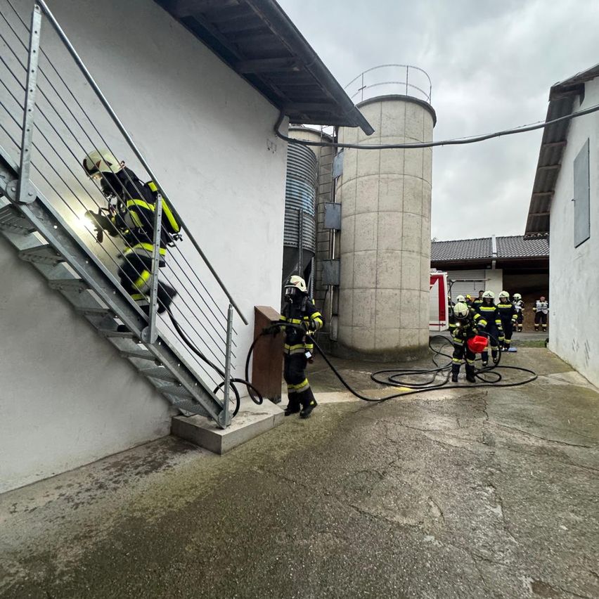 Feuerwehrleute klettern mit einem Seil eine Treppe hinauf und verwenden einen Feuerwehrschlauch in der Nähe eines Gebäudes mit Lagertanks und einem roten Schild.