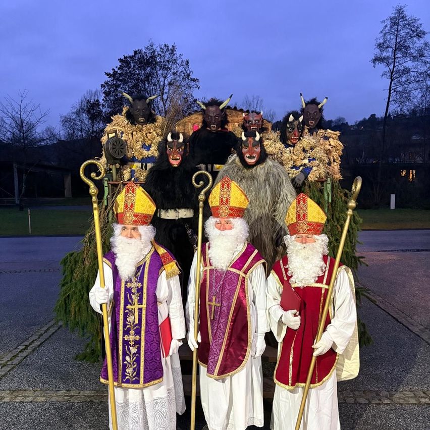 Drei Männer in Bischofskostümen mit weißen und farbigen Gewändern stehen mit Stäben, flankiert von zwei Männern in Teufelskostümen. Bäume und ein Gebäude sind im Hintergrund.