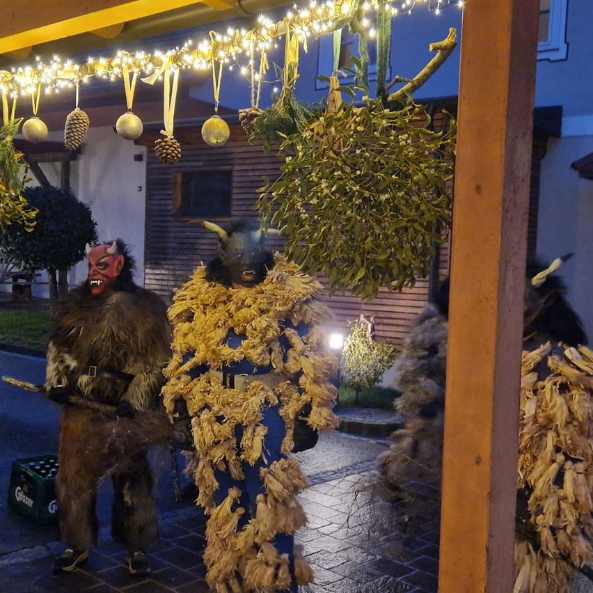 Zwei Personen in Kostümen stehen auf einer Straße, eine trägt eine Fell- und Hornmaske, die andere ein blaues Kostüm mit Heu. Sie stehen unter dekorativen Lichtern und Mistelzweigen.