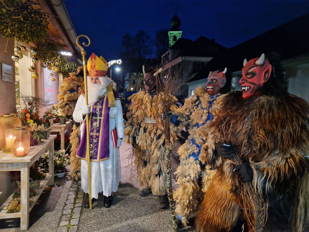 Eine Person als Sankt Nikolaus steht bei Nacht auf der Straße, begleitet von drei Personen in gruseligen Teufelskostümen, die trockene Zweige halten.