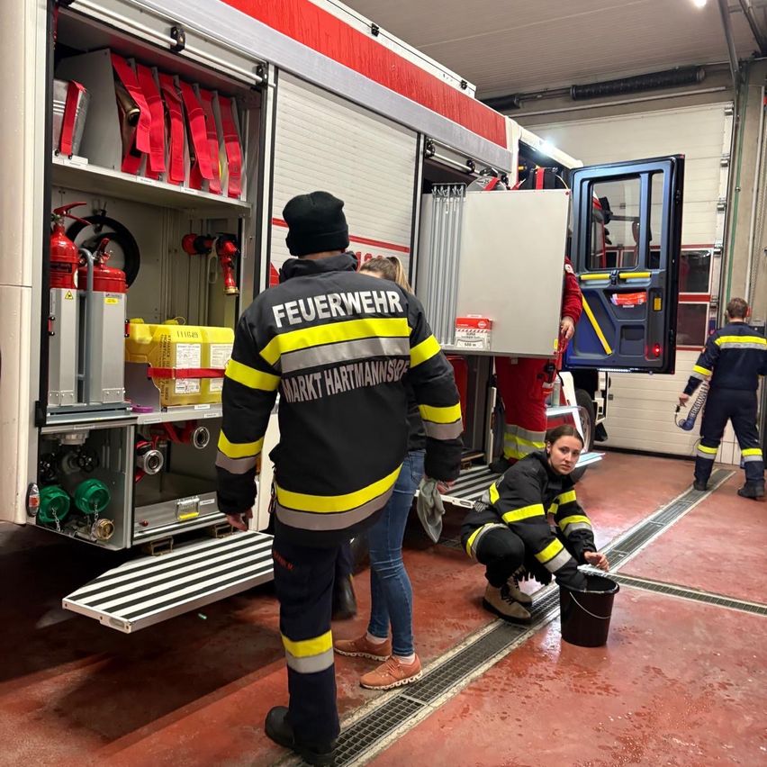Feuerwehrleute bereiten sich in einer Feuerwache auf einen Einsatz vor. Ein Feuerwehrmann in Uniform steht neben dem Feuerwehrwagen, während ein anderer Feuerwehrmann mit einem Eimer kniet.