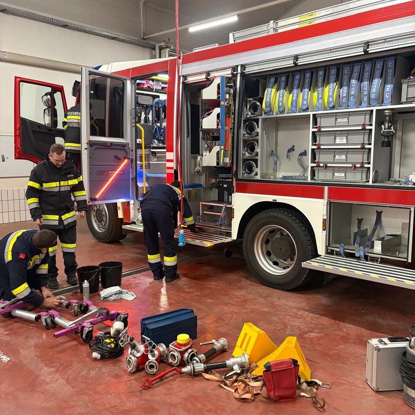 Drei Feuerwehrleute bereiten einen Feuerwehrwagen vor, wobei einer an der Ausrüstung im Inneren arbeitet, während die anderen draußen sind. Das Innere des Wagens ist mit verschiedenen Werkzeugen und Ausrüstungsgegenständen gefüllt.