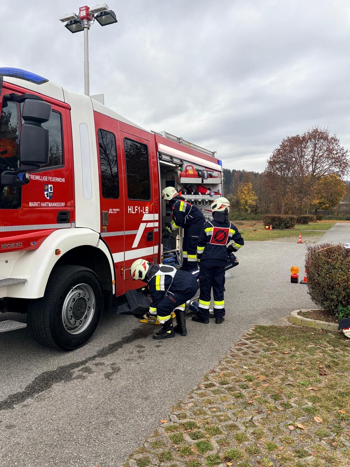 Drei Feuerwehrleute bereiten einen Feuerwehrwagen vor. Einer justiert die Ausrüstung, während ein anderer bereit steht. Die Szene spielt sich auf einem Parkplatz mit Sicherheitskegeln und Bäumen ab.