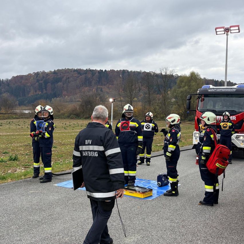 Eine Gruppe von Feuerwehrleuten steht in einer Reihe, trägt Helme und Uniformen, auf einer Betonstraße mit einem Feuerwehrwagen im Hintergrund.