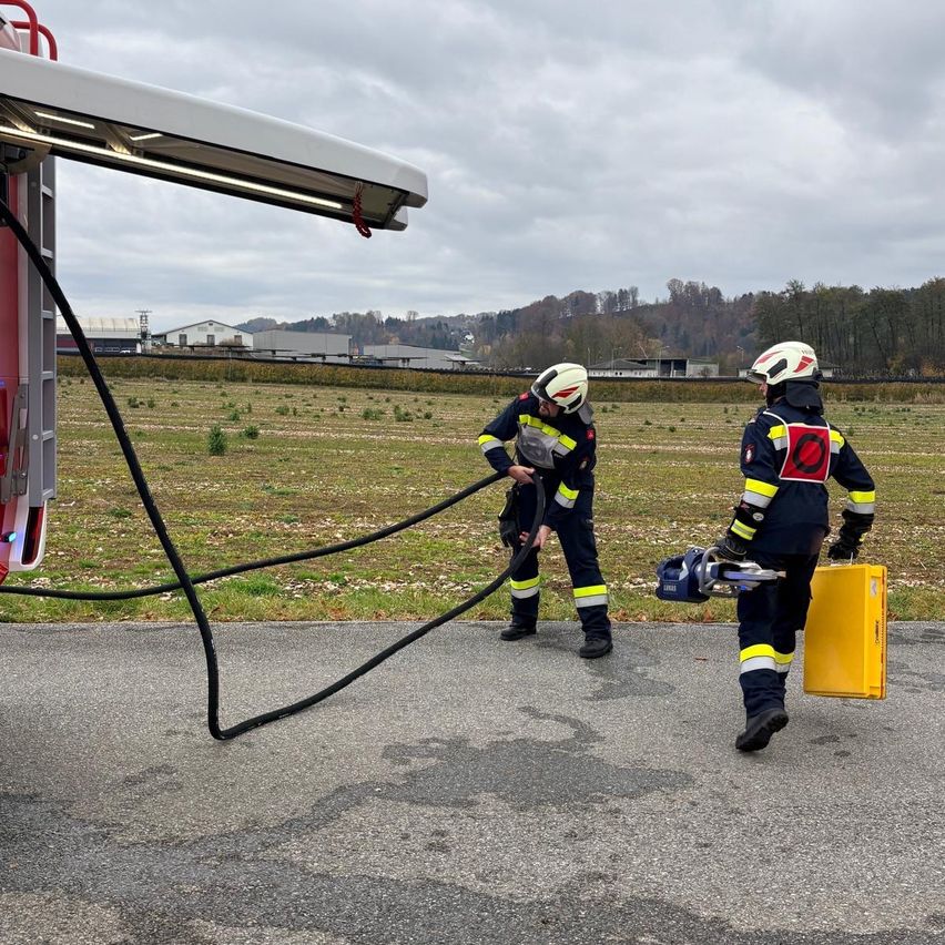 Zwei Feuerwehrleute stehen neben einem Feuerwehrauto, einer hält einen Schlauch und der andere trägt einen gelben Container. Sie befinden sich auf einer asphaltierten Straße mit einem Grasfeld im Hintergrund.