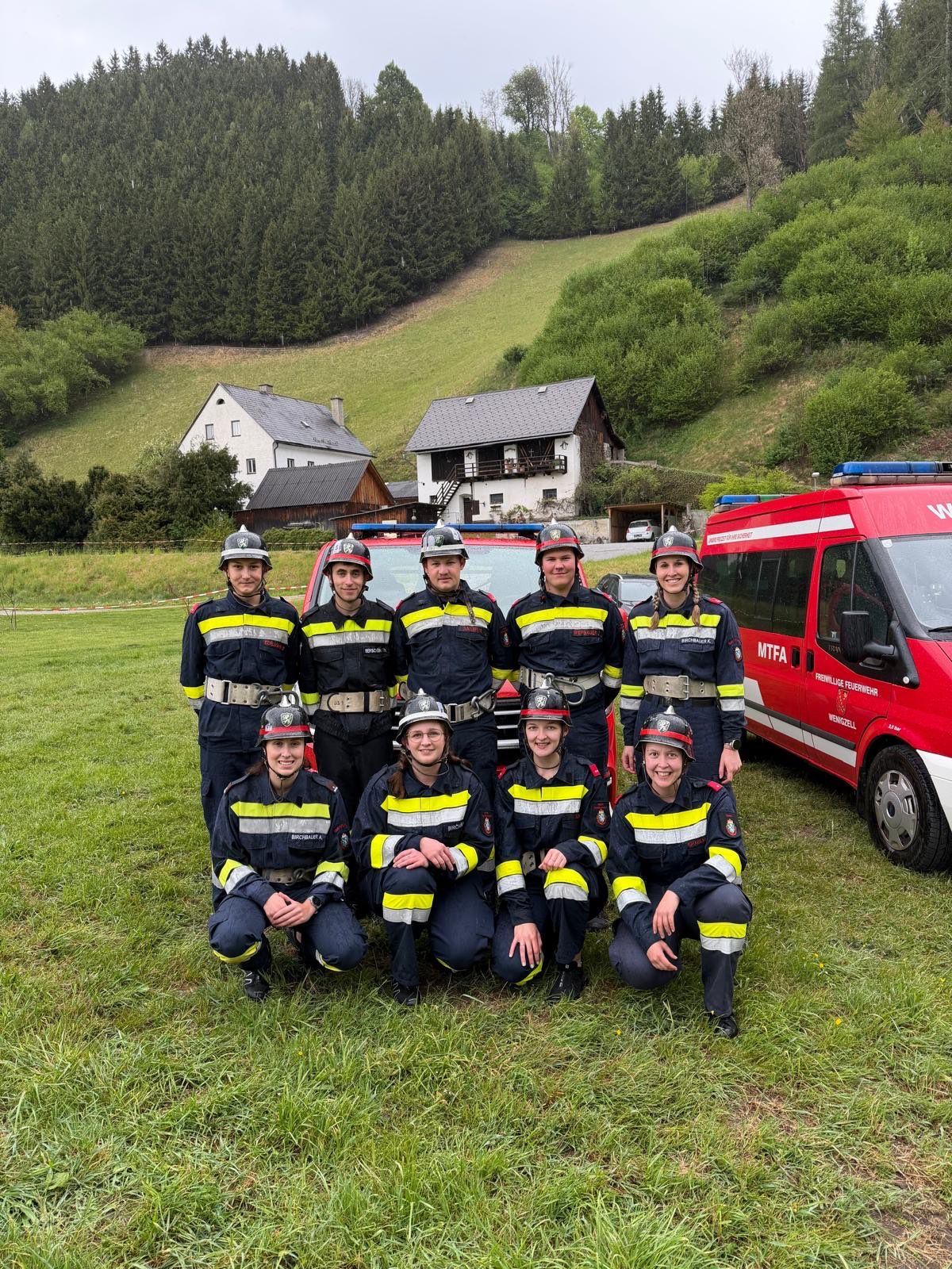 A group of firefighters in uniform poses for a photo in front of a red vehicle and a house on a hill.