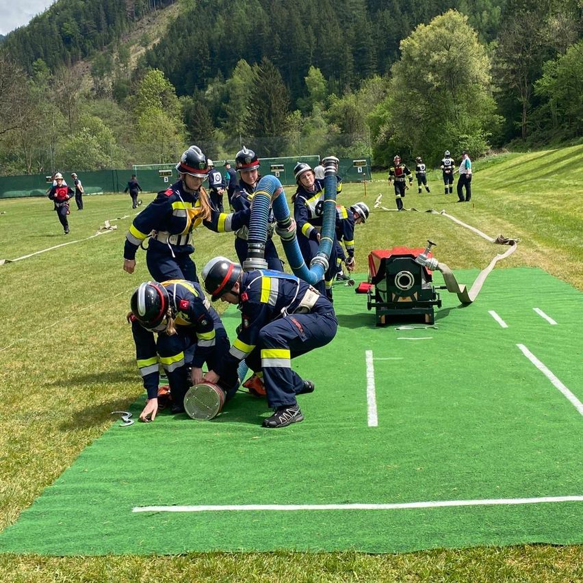 Firefighters are practicing on a green field, with some using a hose while others are working on the ground. Trees and a fence are in the background.