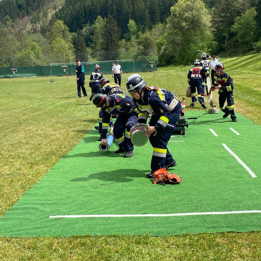 A group of firefighters in uniform, some bent over, are practicing on a green field. They are holding various equipment. Trees and a mountain are in the background.