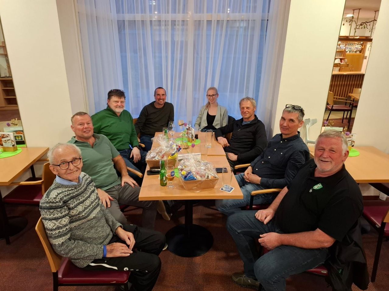 Eine Gruppe lächelnder Menschen sitzt um einen Tisch mit Getränken und Snacks. Der Raum ist hell mit einer weißen Gardine und Regalen im Hintergrund.