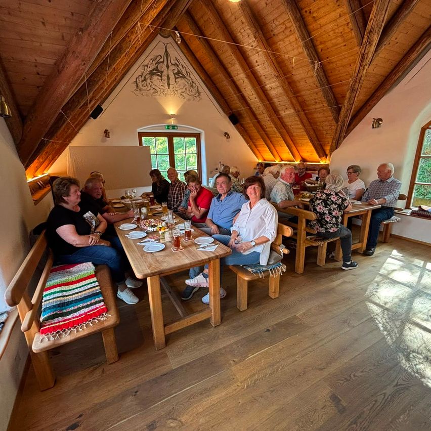 Eine Gruppe von Menschen sitzt um Holztische in einem rustikalen, dachgeschossartigen Essbereich, mit Holzbalken über dem Kopf und Sonnenlicht, das hereinströmt.