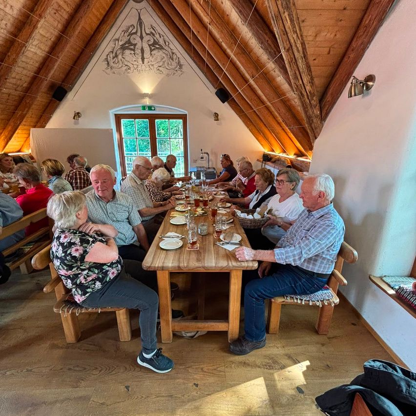 Eine Gruppe älterer Menschen sitzt um einen langen Holztisch in einem rustikalen Raum und genießt ihr Essen. Der Raum hat Holzbalken und eine weiße Wand.