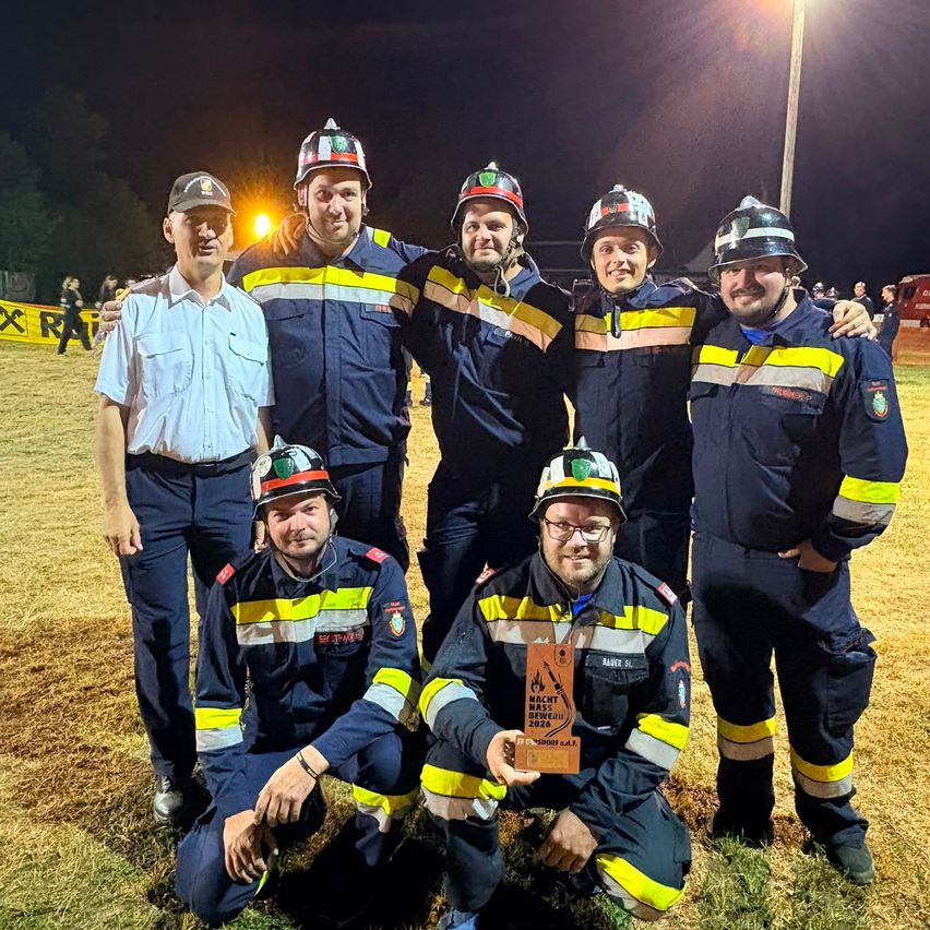 A group of firefighters in uniform are posing for a photo at night. One man is kneeling and holding a trophy.