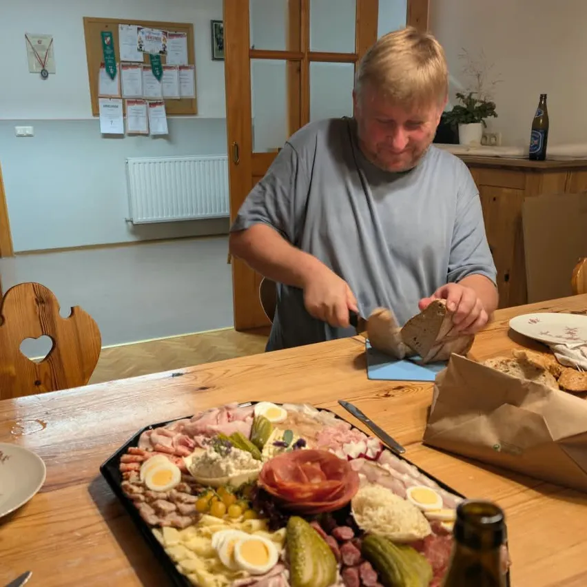 Ein Mann sitzt an einem Tisch und schneidet ein Brot mit einem Messer. Vor ihm steht eine große Platte mit Essen.