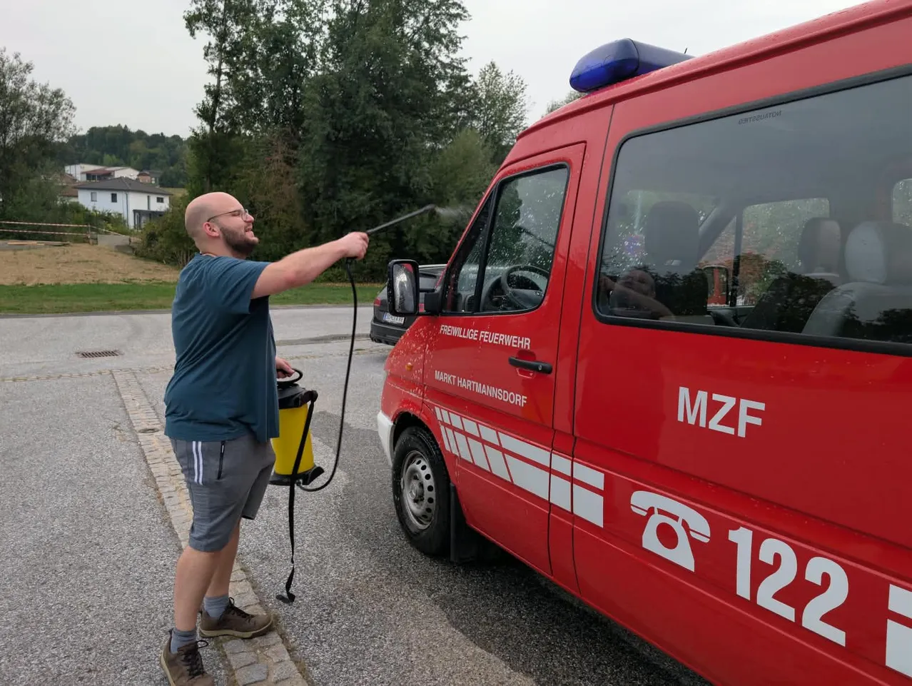 Ein Mann mit Brille und Turnschuhen sprüht Wasser auf einen roten Feuerwehrwagen mit der Aufschrift 'MZF 12' an der Seite.