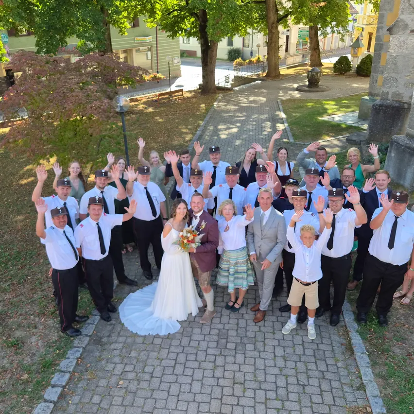 Eine Gruppe von Menschen in formeller Kleidung, einschließlich Braut und Bräutigam, posiert für ein Foto auf einem gepflasterten Weg in einem Park.