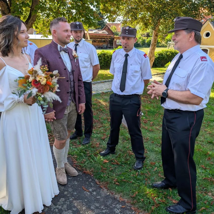 Eine Braut und ein Bräutigam stehen mit zwei uniformierten Männern in einem Park, einer hält einen Blumenstrauß. Der Bräutigam trägt eine Fliege und Shorts.