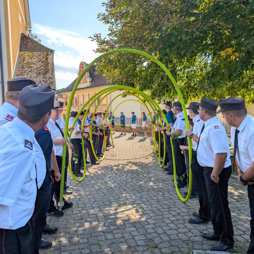 Eine Gruppe uniformierter Personen steht in einer Reihe, hält grüne Bögen, mit einem Gebäude und Personen im Hintergrund an einem sonnigen Tag.