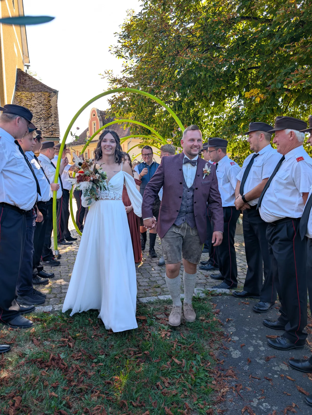 Ein frisch verheiratetes Paar geht Hand in Hand lächelnd einen Weg entlang, der von Männern in traditionellen Uniformen gesäumt ist. Die Braut trägt ein weißes Kleid und einen Blumenstrauß. Der Bräutigam ist in einem Anzug und traditioneller bayerischer Kleidung gekleidet.