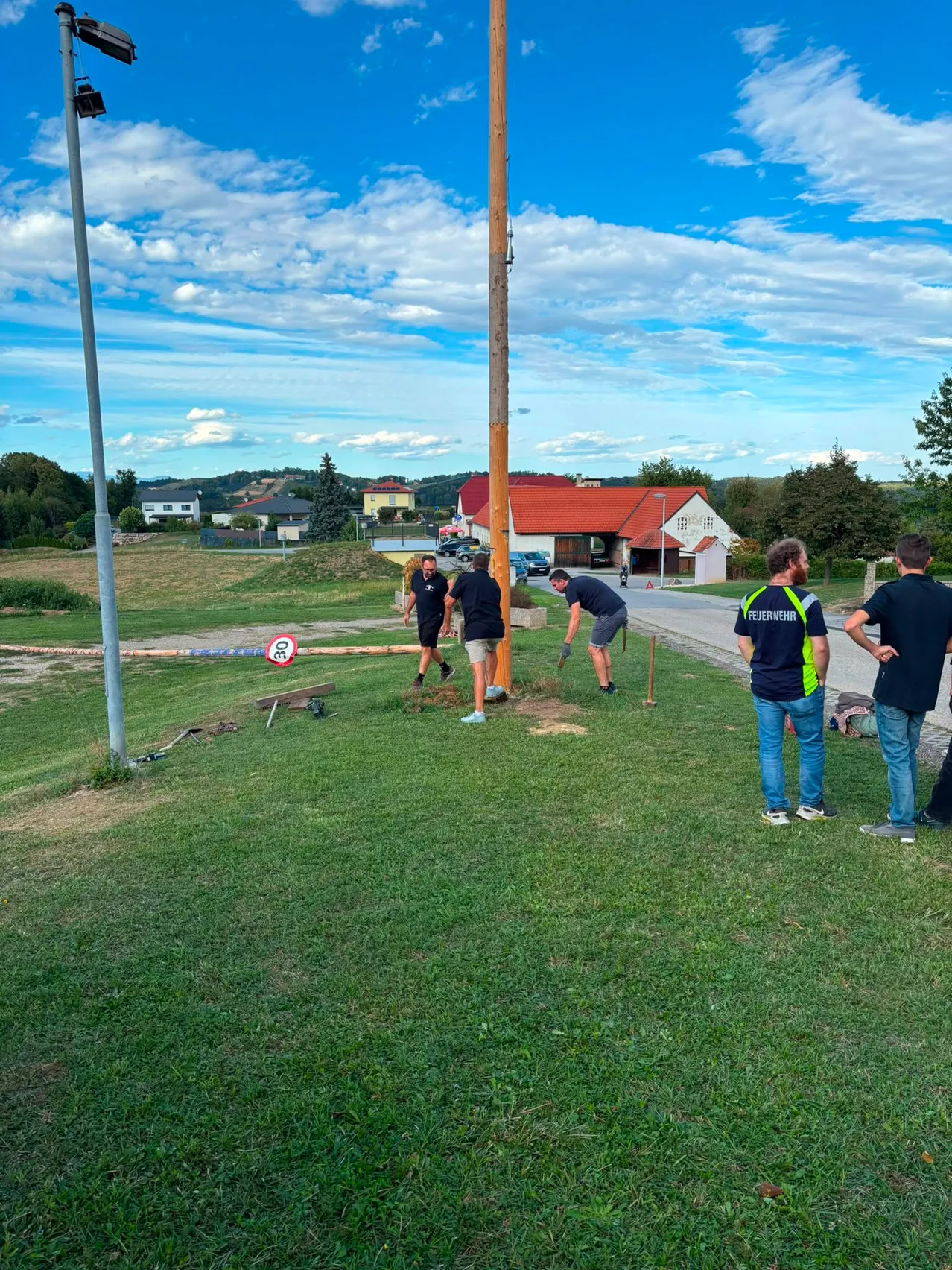 Fünf Männer arbeiten auf einem Rasenfeld, zwei beugen sich vor und drei stehen. Ein Pfahl steht in der Mitte und ein Schild liegt auf dem Gras. Gebäude und Bäume sind im Hintergrund.
