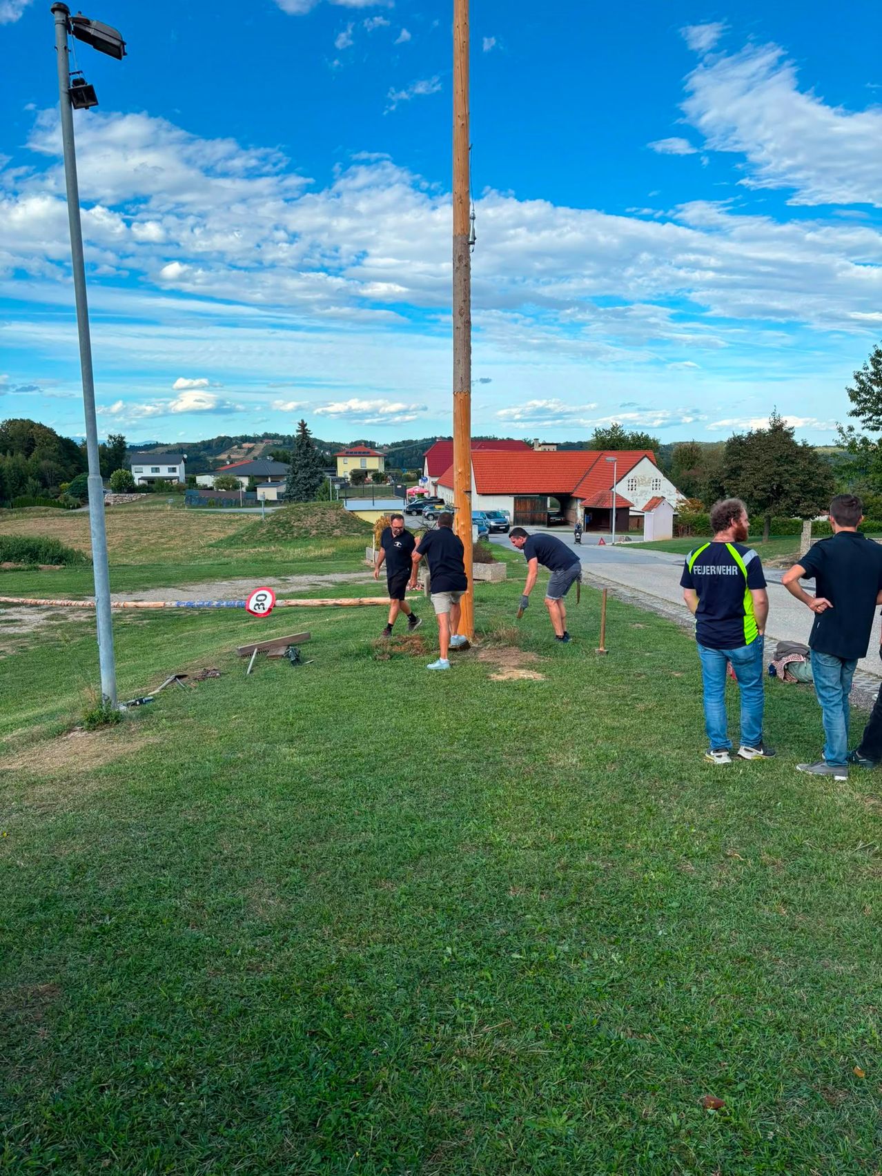 Five men are working on a grassy field, with two bending over and three standing. A pole is in the middle, and a sign is on the grass. Buildings and trees are in the background.