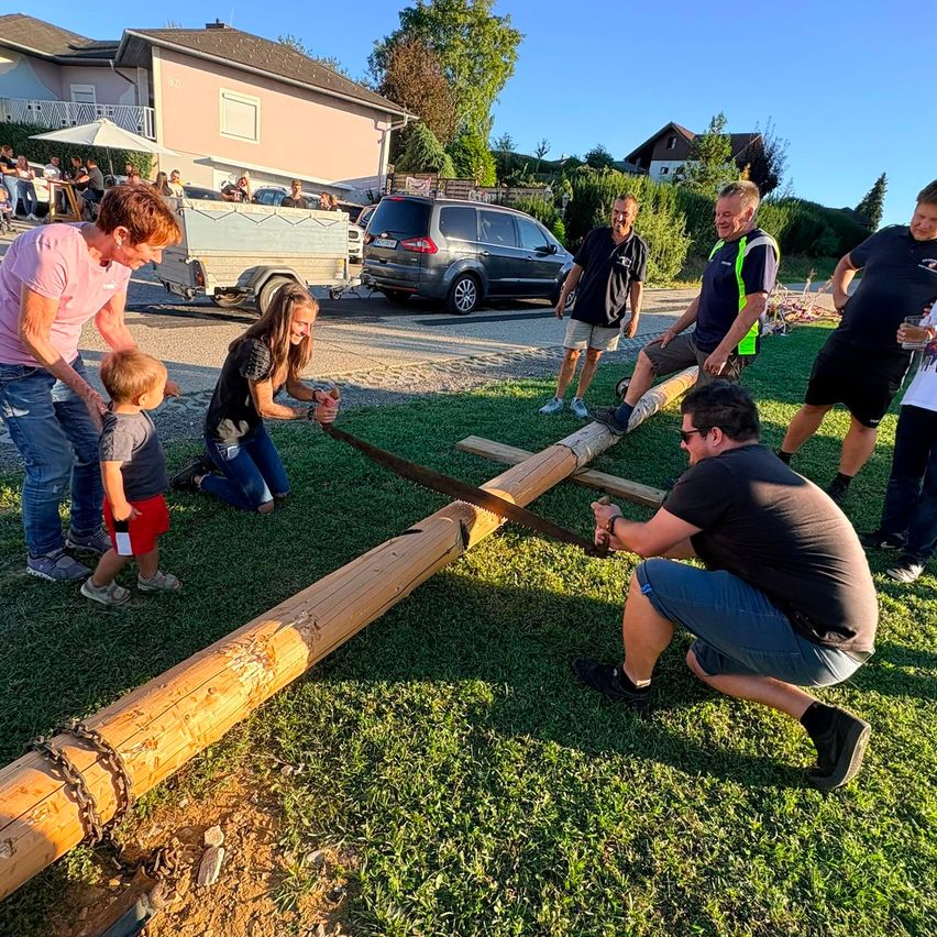 A group of people are gathered on a lawn, using a large saw to cut a wooden log. Some are kneeling, others standing, with a child holding a small saw. A house and cars are in the background.