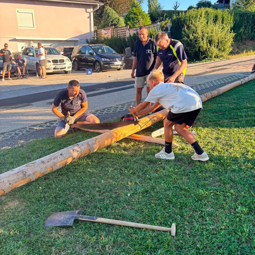A group of men are working on a large log on the grass, with one using a saw and another holding a shovel. Behind them, several cars are parked, and people are standing near a building.