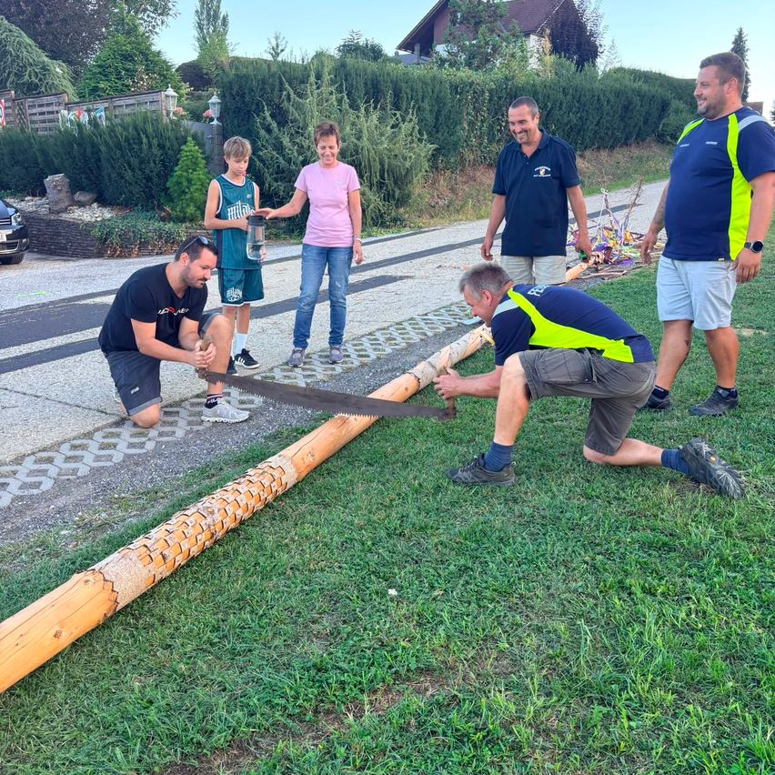 Five people are working on a large wooden log on a grassy area. Two men are kneeling and sawing the log while others watch.