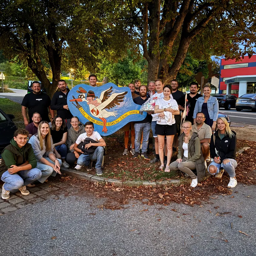 Eine Gruppe von Menschen posiert für ein Foto vor einem großen Storchschild mit Datum und Name.