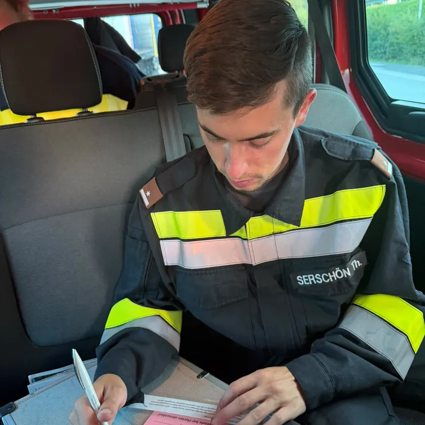 Ein Mann in einer reflektierenden Sicherheitsjacke sitzt im Rücksitz eines Fahrzeugs und schreibt auf einem rosa Formular mit einem Stift.