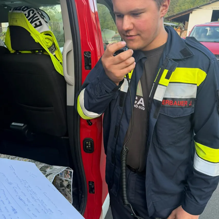 Ein Feuerwehrmann in einem blau-gelben Jackett steht neben einem roten Fahrzeug und hält ein Funkgerät an sein Ohr.