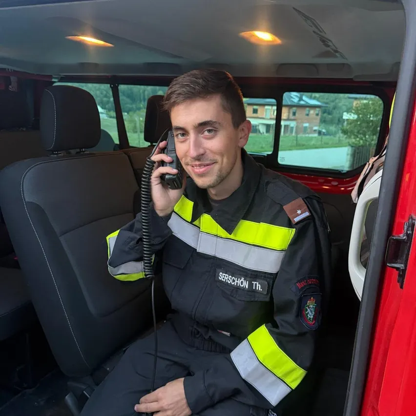 Ein Feuerwehrmann in einem roten Fahrzeug, der eine reflektierende Weste trägt, sitzt und telefoniert. Das Fahrzeug hat ein schwarzes Interieur mit sichtbaren Sitzen und einem Fenster, das ein Haus und Bäume zeigt.