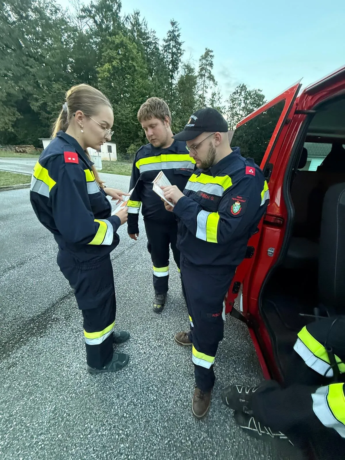 Drei Feuerwehrleute in Uniform stehen neben einem roten Fahrzeug, einer liest ein Dokument, während die anderen zusehen.