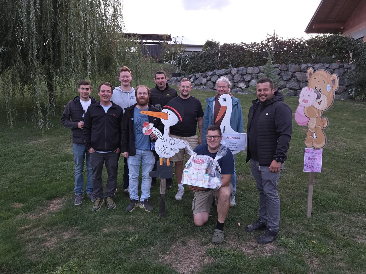 Eine Gruppe von Männern steht zusammen auf einem Rasenfeld, einige halten eine Storch-Ausschnitt und ein Babygeschenk. Sie lächeln und posieren für ein Foto.