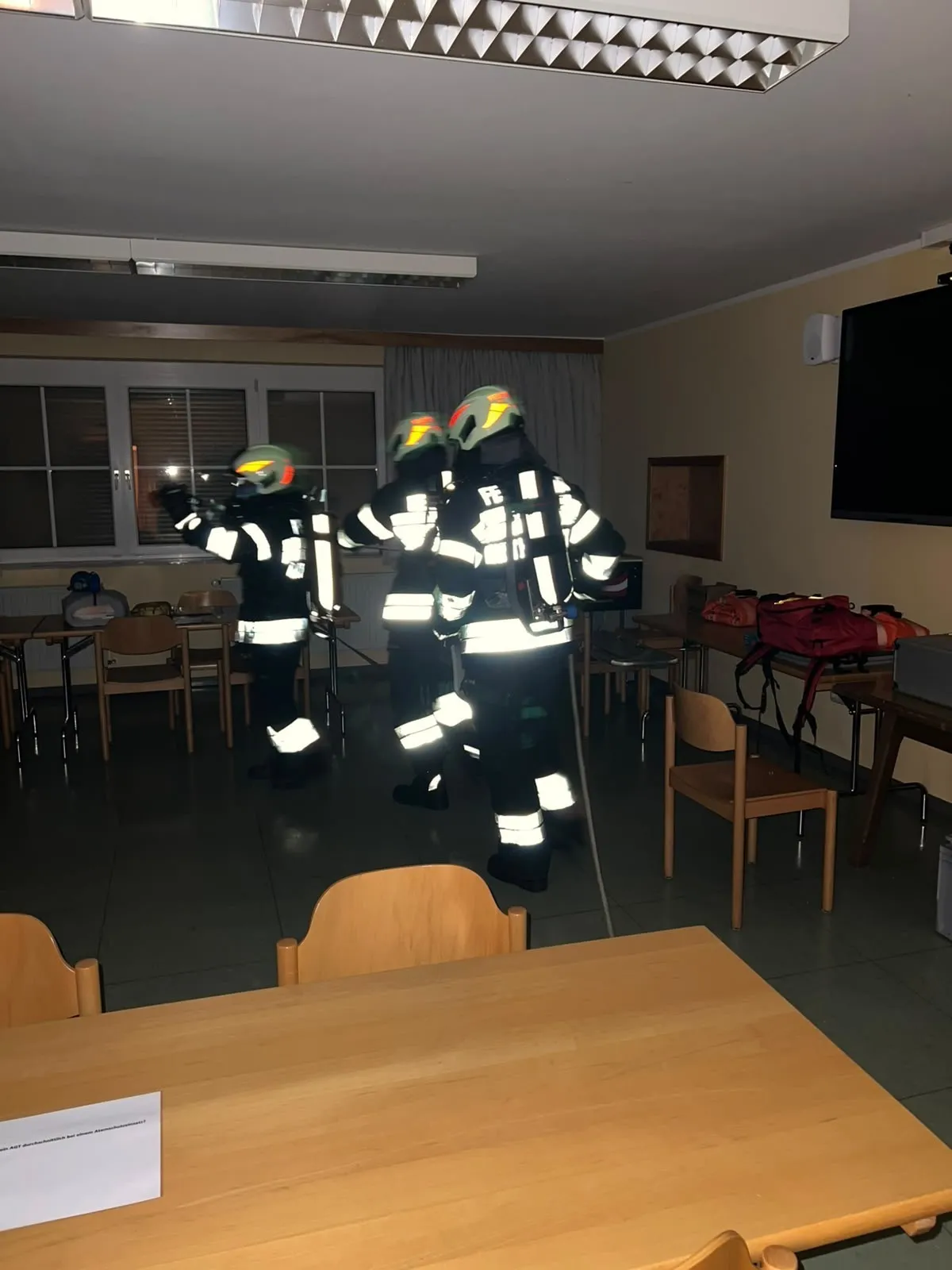 Drei Feuerwehrleute in voller Ausrüstung trainieren in einem schwach beleuchteten Raum mit Stühlen und Tischen.