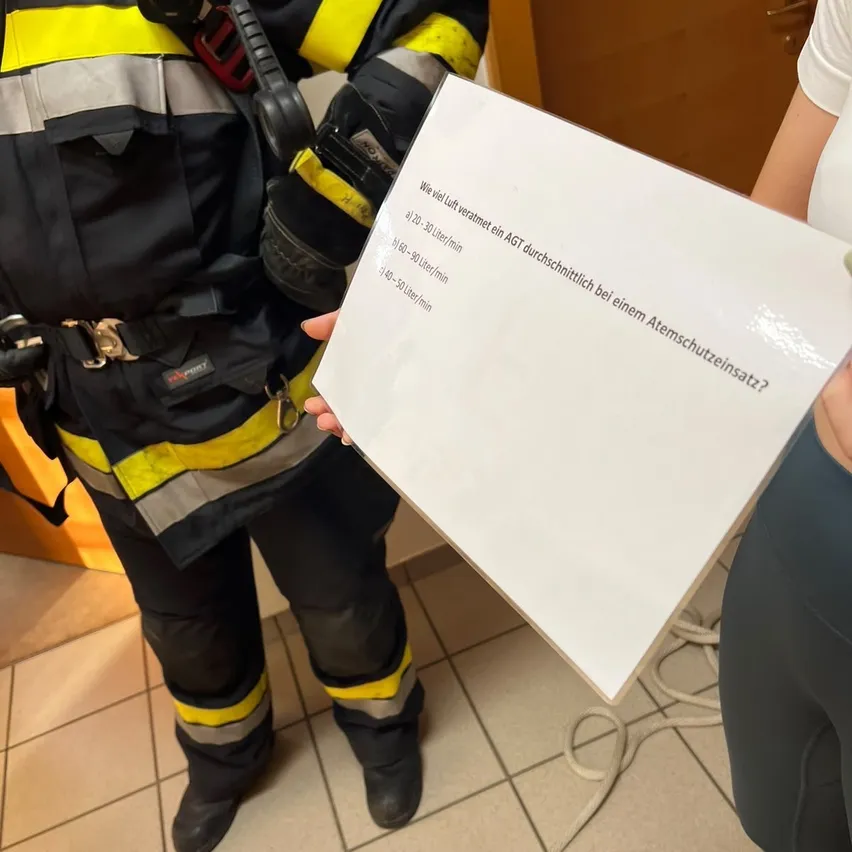 Ein Feuerwehrmann in voller Ausrüstung hält ein weißes Blatt Papier mit Anweisungen. Der Text ist auf Deutsch. Der Feuerwehrmann steht in einem gefliesten Raum.