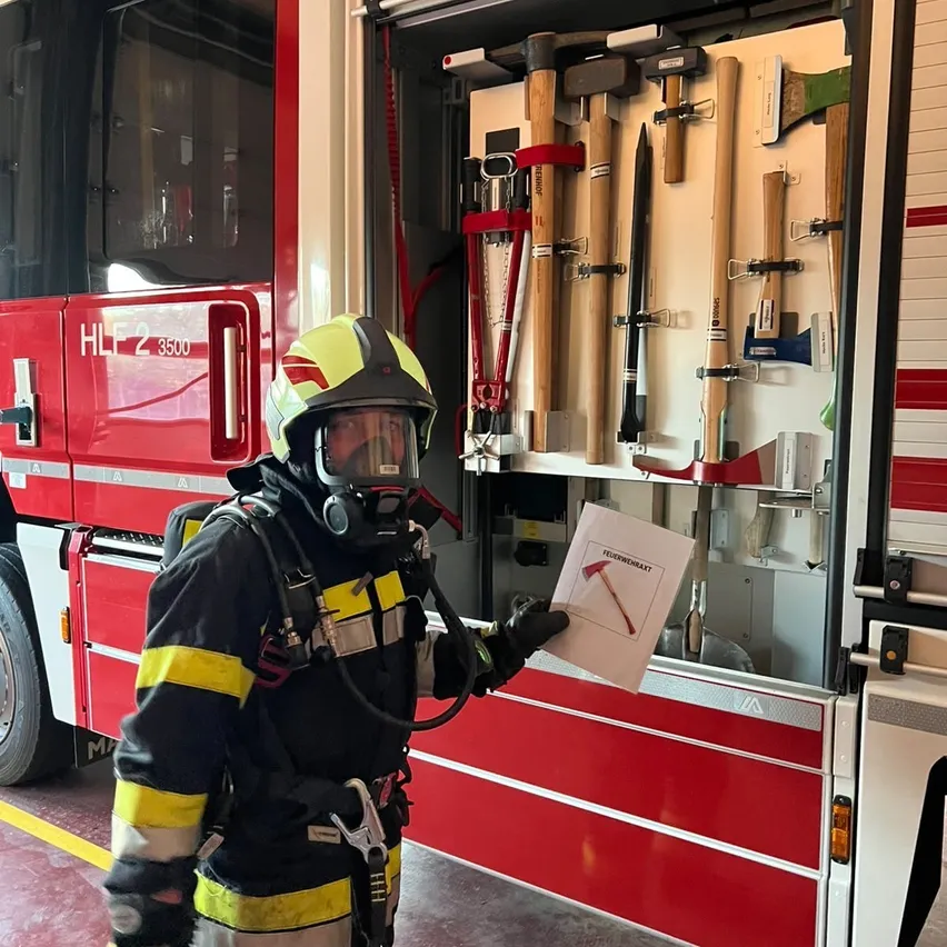 Ein Feuerwehrmann in voller Ausrüstung steht neben einem Feuerwehrauto und hält ein Papier mit einer Hammerzeichnung in der Hand.