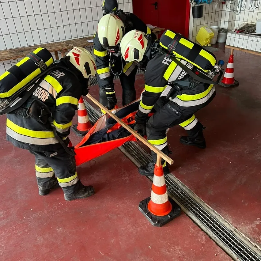 Vier Feuerwehrleute in voller Ausrüstung üben in einer Trainingseinrichtung eine Rettungsaktion. Eine Person liegt auf einer Trage, während die anderen sie hochheben.