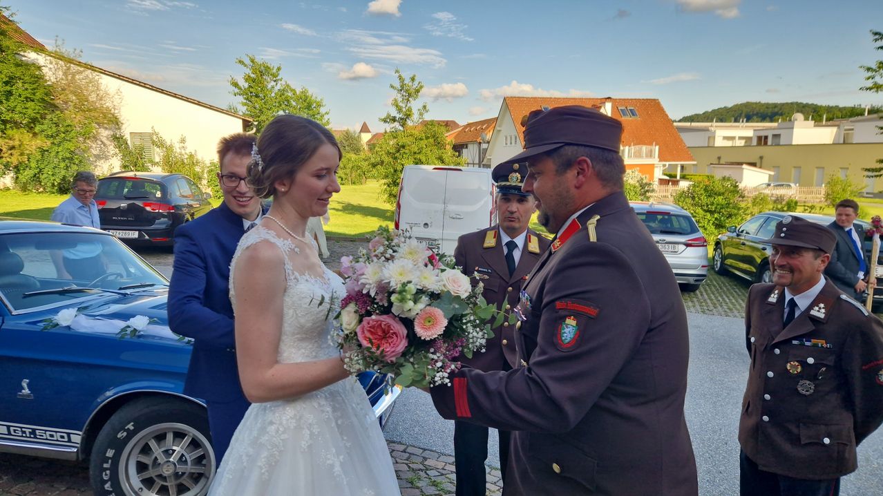 A bride in a white dress holding a flower bouquet looks at a man in uniform who is offering the flowers. Behind them is a man in a suit and glasses.